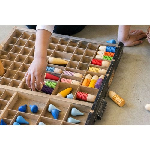 Een hand sorteert grapat houten brots poppetjes en blauwe vormen in een houten vakkenbak.