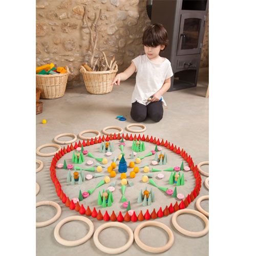Een kind speelt met Grapat houten kegels en ringen, gerangschikt in een kleurrijk mandala-patroon op de vloer.