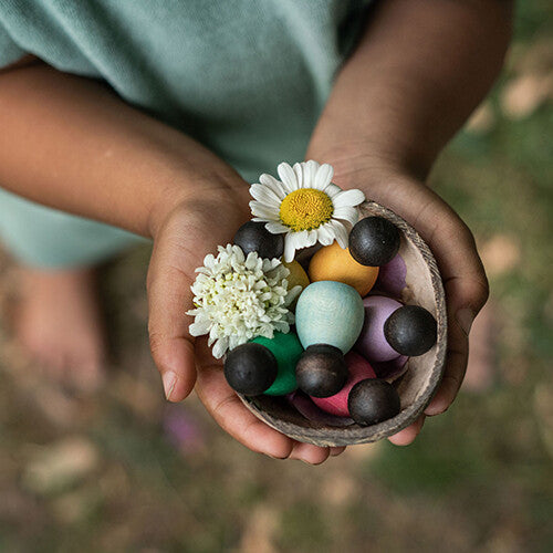 Kinderhanden houden een kokosnootkom vast met grapat houten nins® poppetjes baby's, donkere ballen en bloemen.