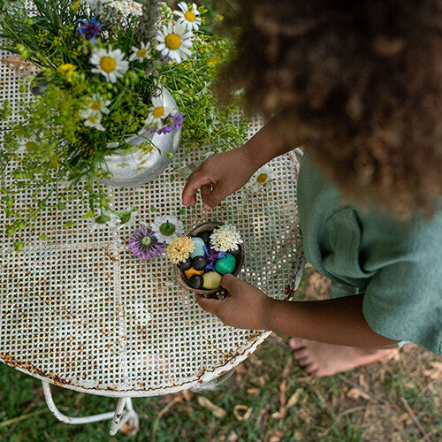 Een kind speelt met grapat houten nins® poppetjes baby's en bloemen op een witte tuintafel.