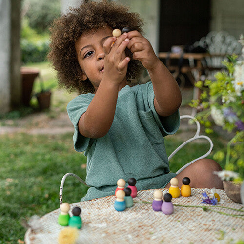 Een kind met krullend haar speelt buiten aan een tafel met grapat houten nins® poppetjes baby's.