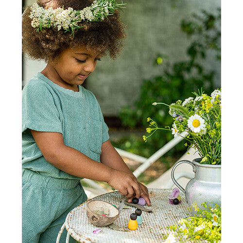 Een kind met een bloemenkrans speelt met grapat houten nins® poppetjes baby's op een witte tuintafel.