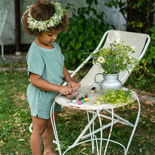 Een kind met een bloemenkrans speelt met grapat houten nins® poppetjes baby's aan een witte tuintafel in de tuin.