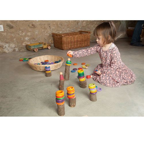 Een peuter speelt op de grond met Grapat houten Nins poppetjes, ringen en schijfjes, gestapeld op houten stammetjes.