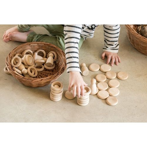 Een kind speelt op de grond met Grapat houten Nins® poppetjes, ringen en schijfjes als stapelspeelgoed, naast een mand vol houten speelgoed.