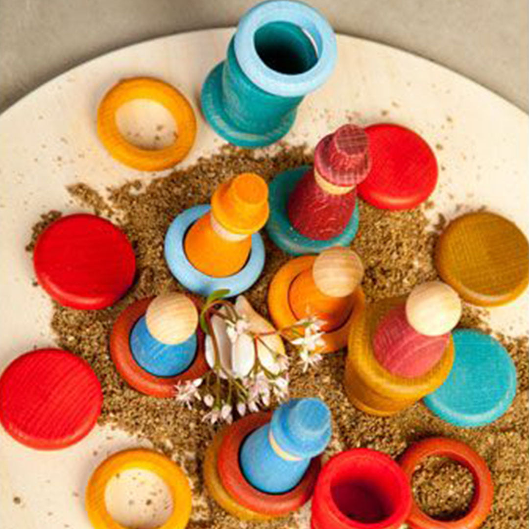 Grapat houten nins® poppetjes met ringen en schijfjes liggen verspreid op een ronde ondergrond met zand en bloemetjes.