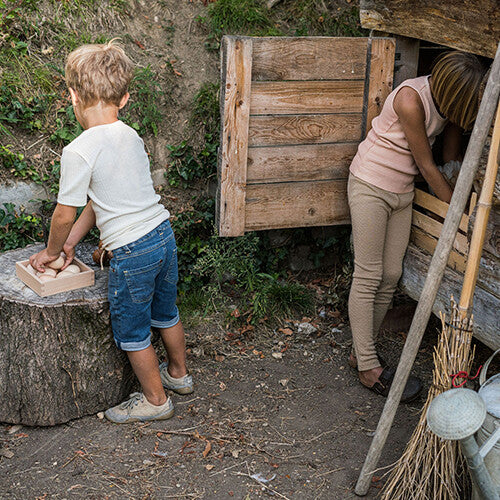 Twee kinderen spelen buiten; een jongen legt houten speelgoed eieren in een Grapat houten opbergbak op een boomstronk.