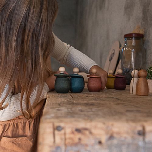 Een kind reikt naar een rij grapat houten poppetjes in bakjes op een houten tafel.