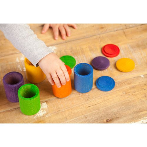 Een kinderhand pakt een oranje houten potje van het kleurrijke sorteerspeelgoed op een houten ondergrond.