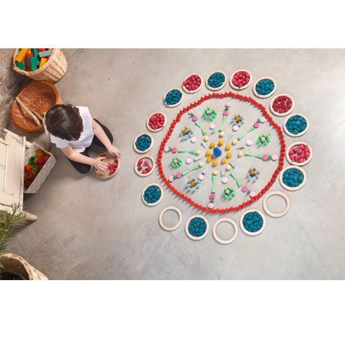 Een kind speelt op de grond met Grapat houten speelgoed, waaronder blauwe schijfjes, gerangschikt in een kleurrijke mandala.