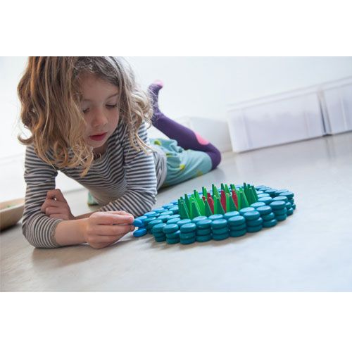 Een kind ligt op de grond en speelt met Grapat bouwspeelgoed, bestaande uit blauwe houten schijfjes en groene staafjes.