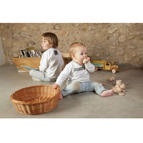 Een baby zit op de grond en stopt een houten vorm in de mond, met een rieten mand en andere houten vormen ernaast.