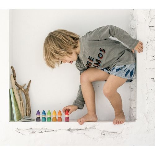 Een kind speelt met een rij grapat houten kabouter speelgoedfiguurtjes in regenboogkleuren op een witte vensterbank.