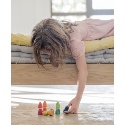Een kind ligt op bed en reikt naar grapat houten kabouter speelgoedfiguurtjes in regenboogkleuren op de vloer.