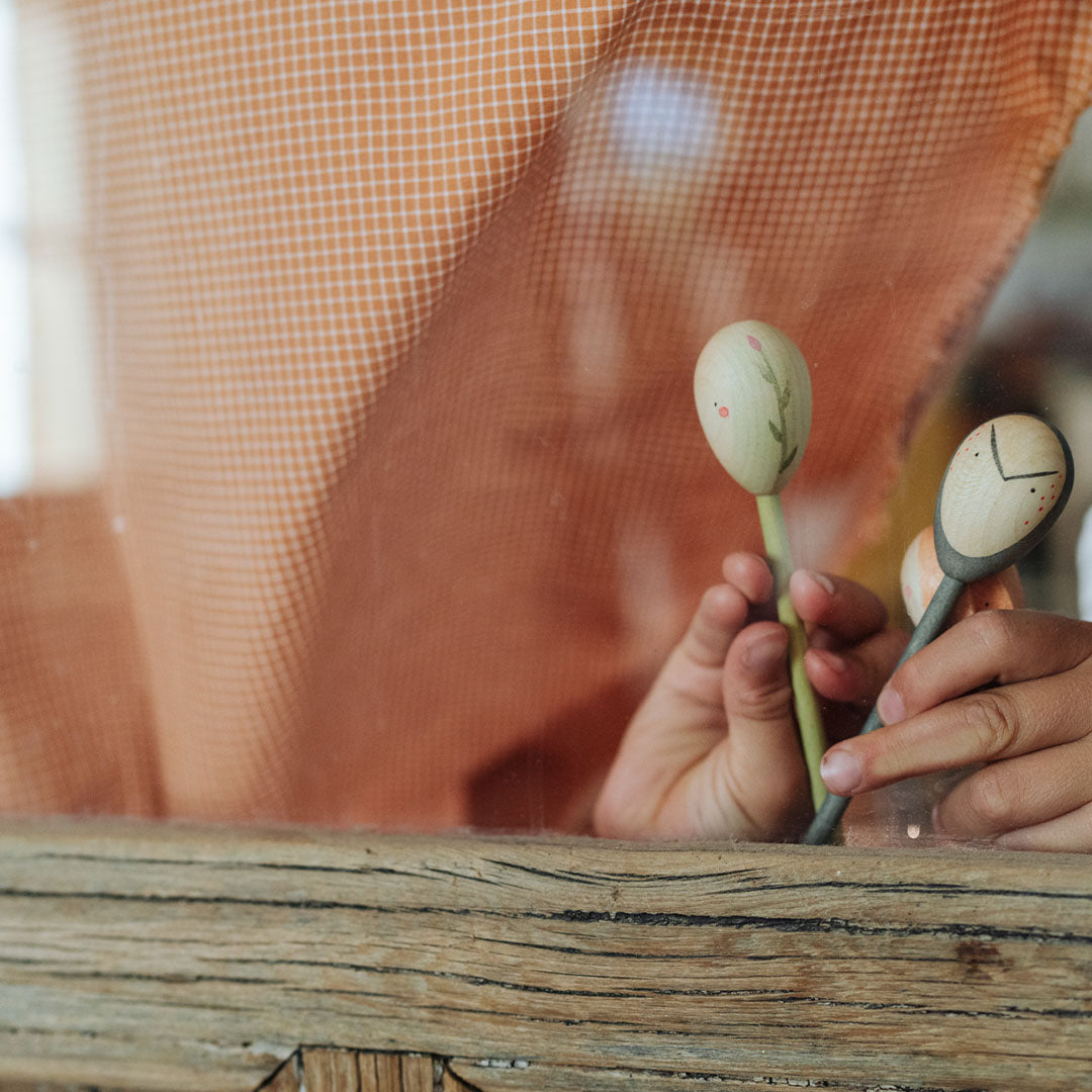Een kind houdt twee Grapat houten speelfiguren met plant- en figuurgezichten vast bij een raam.