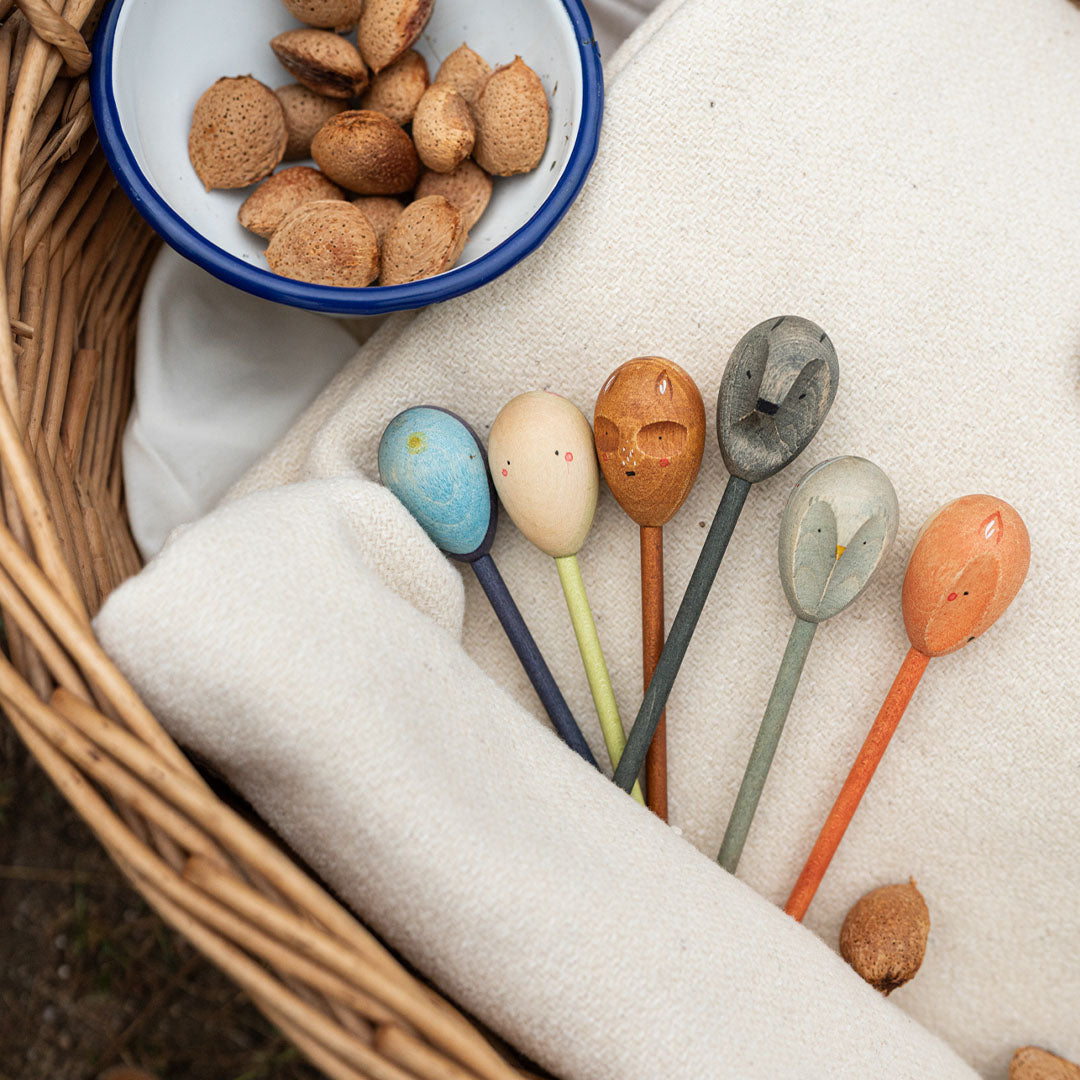 Een collectie Grapat houten speelfiguren met dieren- en figuurgezichten in een rieten mand met een witte doek en amandelen.