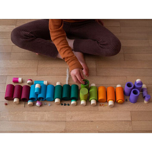 Een kind speelt met Grapat houten cilindervormige blokken in regenboogkleuren, gesorteerd op een houten vloer.