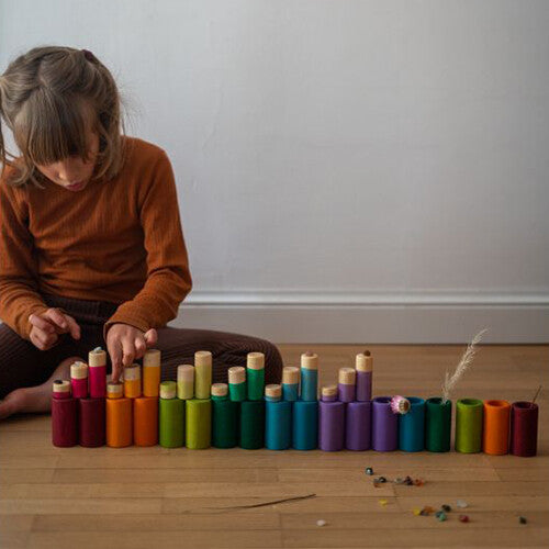 Een kind speelt met Grapat houten cilindervormige blokken in regenboogkleuren, opgesteld als een stad op een houten vloer.