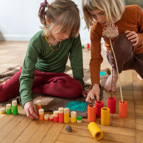 Twee kinderen spelen op een kleed met een grote verzameling grapat houten poppetjes en buizen in diverse kleuren.