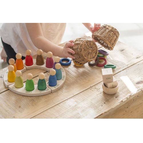 Een kind speelt met een Grapat houten jaarkalender met gekleurde peg dolls en ringen, en kleine rieten mandjes.