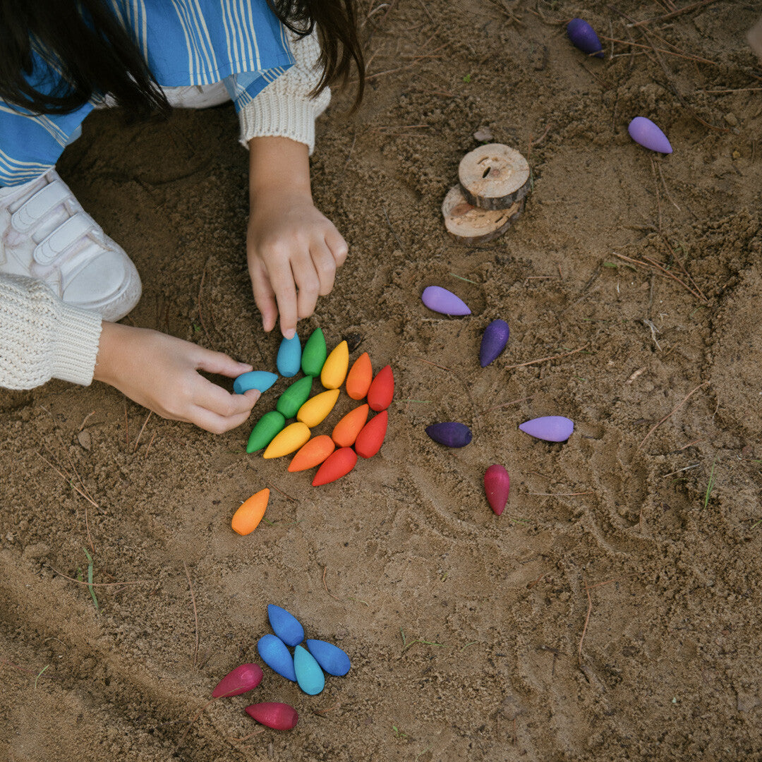 Bovenafzicht van kinderhanden die Grapat houten sneeuwvlokken in regenboogkleuren op zand rangschikken.