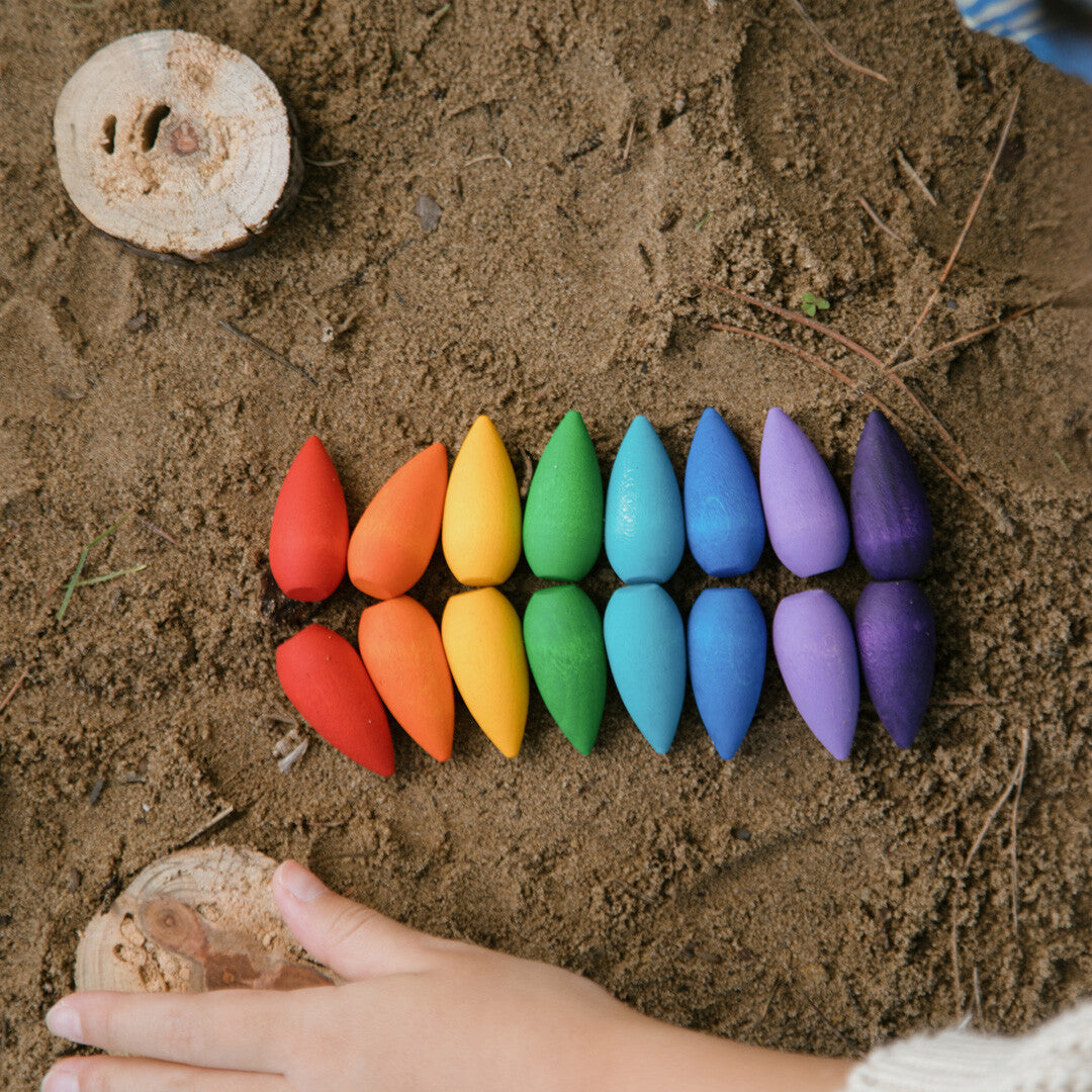 Bovenafzicht van een kinderhand naast Grapat houten sneeuwvlokken in regenboogkleuren, gerangschikt op zand.