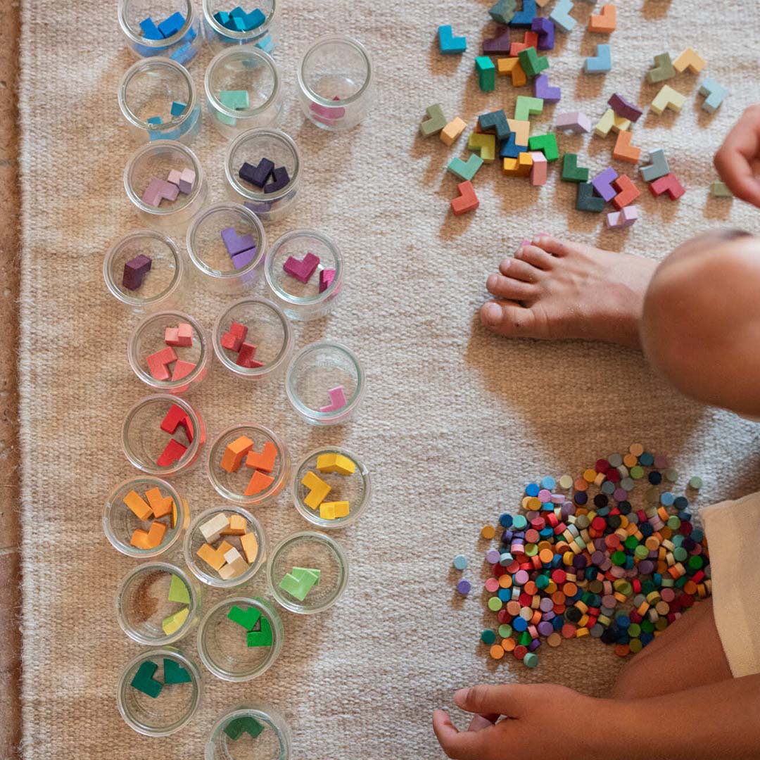 Een kind sorteert Grapat houten sorteervormpjes op kleur in kleine glazen potjes op een vloerkleed.