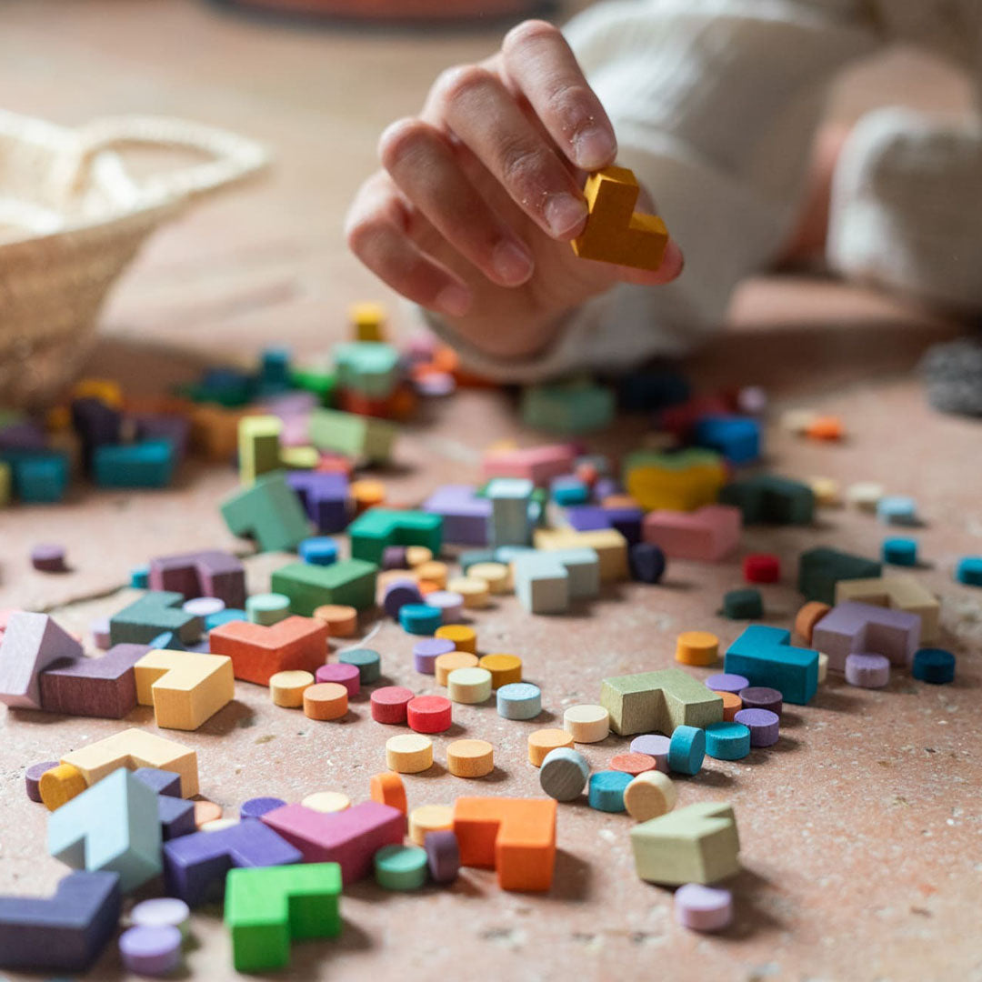 Een kinderhand pakt een geel houten sorteervormpje van de kleurrijke Grapat educatieve speelgoedstukken op de vloer.