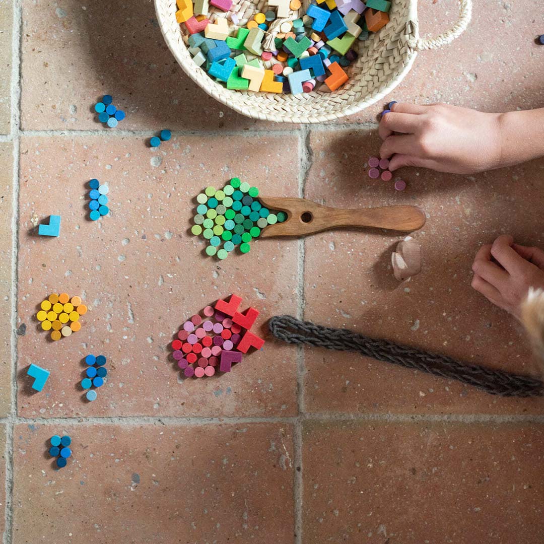 Een kind speelt met Grapat houten sorteervormpjes op een tegelvloer, met een mand vol speelgoed en vormen gesorteerd met een houten schep.