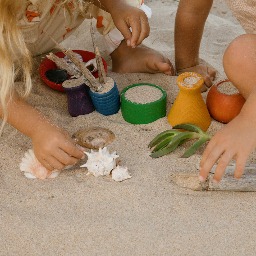 Twee kinderen spelen in het zand met diverse grapat houten speelbakjes gevuld met zand, schelpen en takjes.