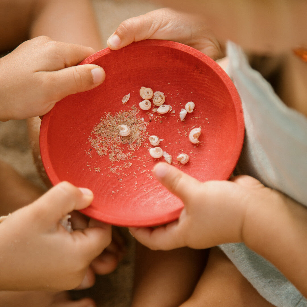 Kinderhanden houden een rood grapat houten speelbakje vast met kleine schelpjes en zand erin.