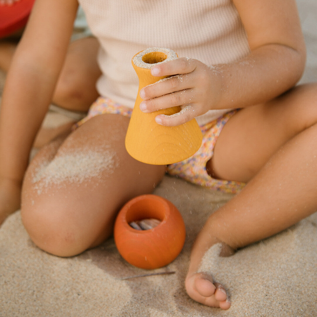 Een kind zit in het zand en houdt een geel grapat houten speelbakje vast waar zand uit stroomt.