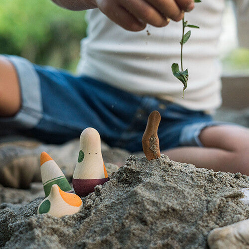 Een kind speelt in het zand met drie grapat houten speelfiguren, waarvan één in een zandhoop staat.