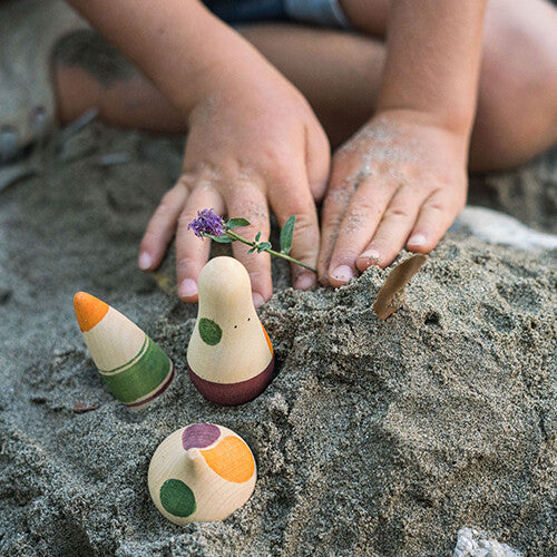 Kinderhanden spelen in het zand met drie grapat houten speelfiguren en een paarse bloem.