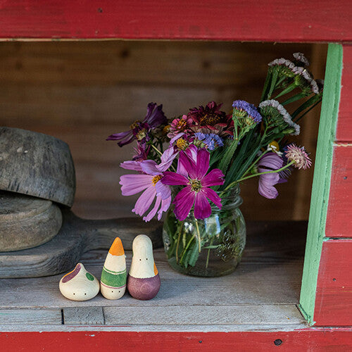 Drie grapat houten speelfiguren staan naast een glazen pot met kleurrijke bloemen op een houten plank in een rood kastje.