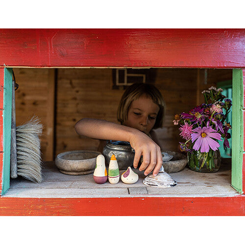 Een kind speelt met de grapat houten speelfiguren en andere voorwerpen op een houten plank in een rood speelhuisje.