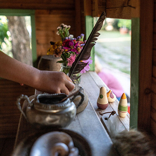 Een kinderhand pakt een veer uit een pot, met de grapat houten speelfiguren op de achtergrond in een speelhuisje.