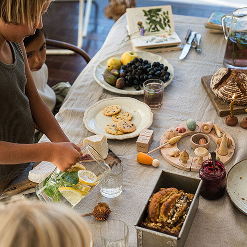 Een kind schenkt water in een glas aan een tafel met eten en een Grapat houten jaarring met speelfiguren wonders.