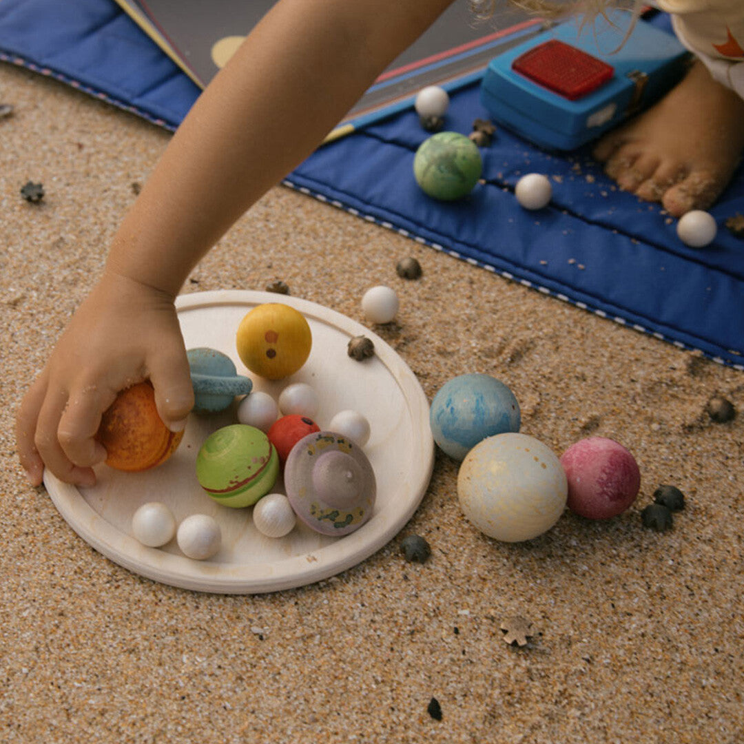 Een kinderhand pakt een kleurrijke houten planeet van een ronde schaal met Grapat sterrenstelsel speelgoed op zand.