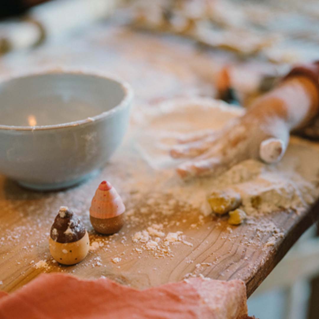 Twee Grapat houten speelgoed bakwaren liggen op een bebloemde houten tafel, met een kom en kinderhand in de achtergrond.