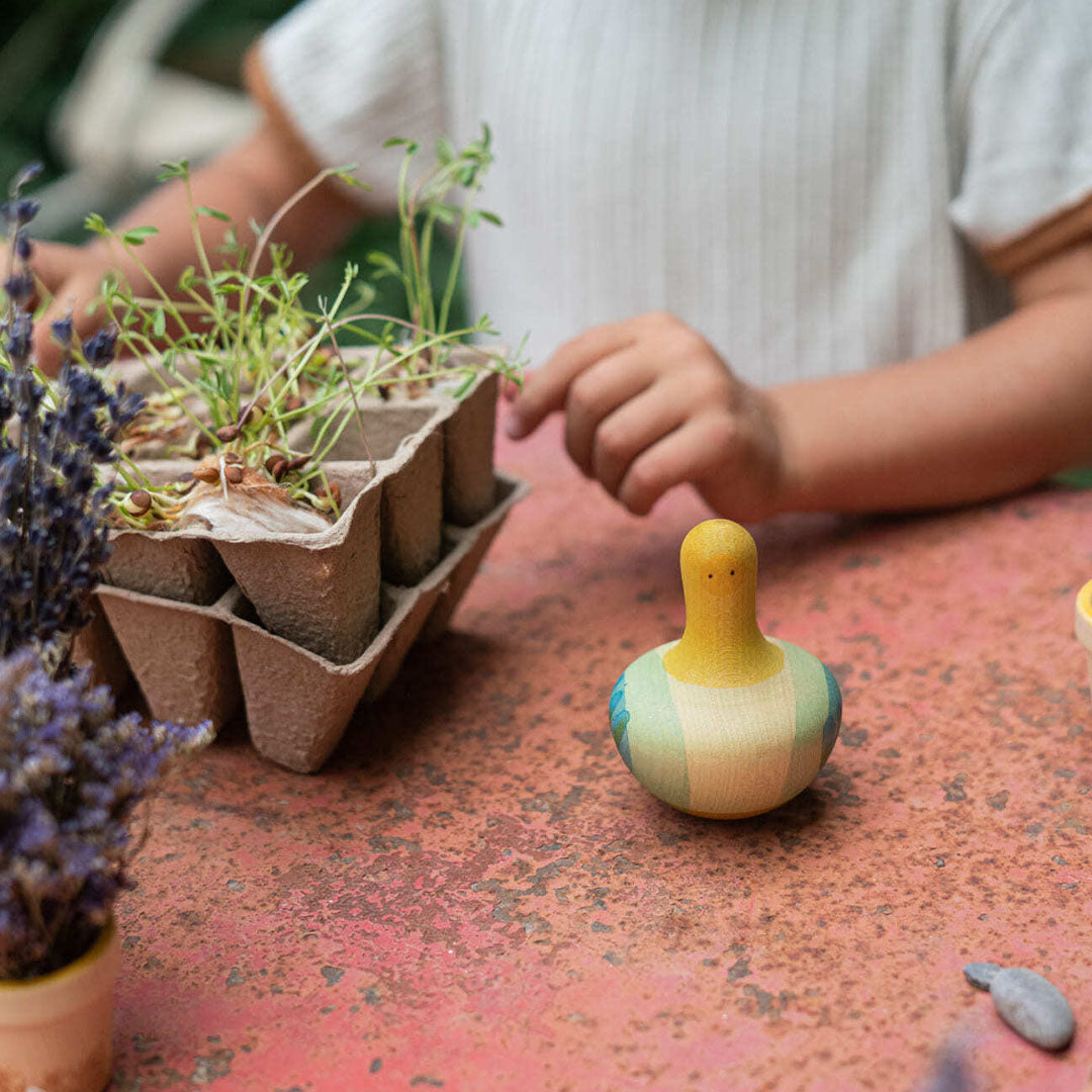 Een kind speelt met de Grapat houten tuimelaar en tol vogel op een tafel naast plantjes.