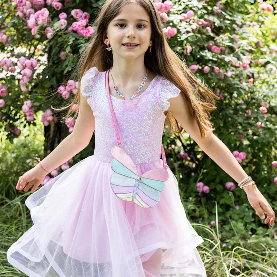 Meisje draagt de Great Pretenders roze pailletten feestjurk met een vlindertasje in een tuin met roze bloemen.