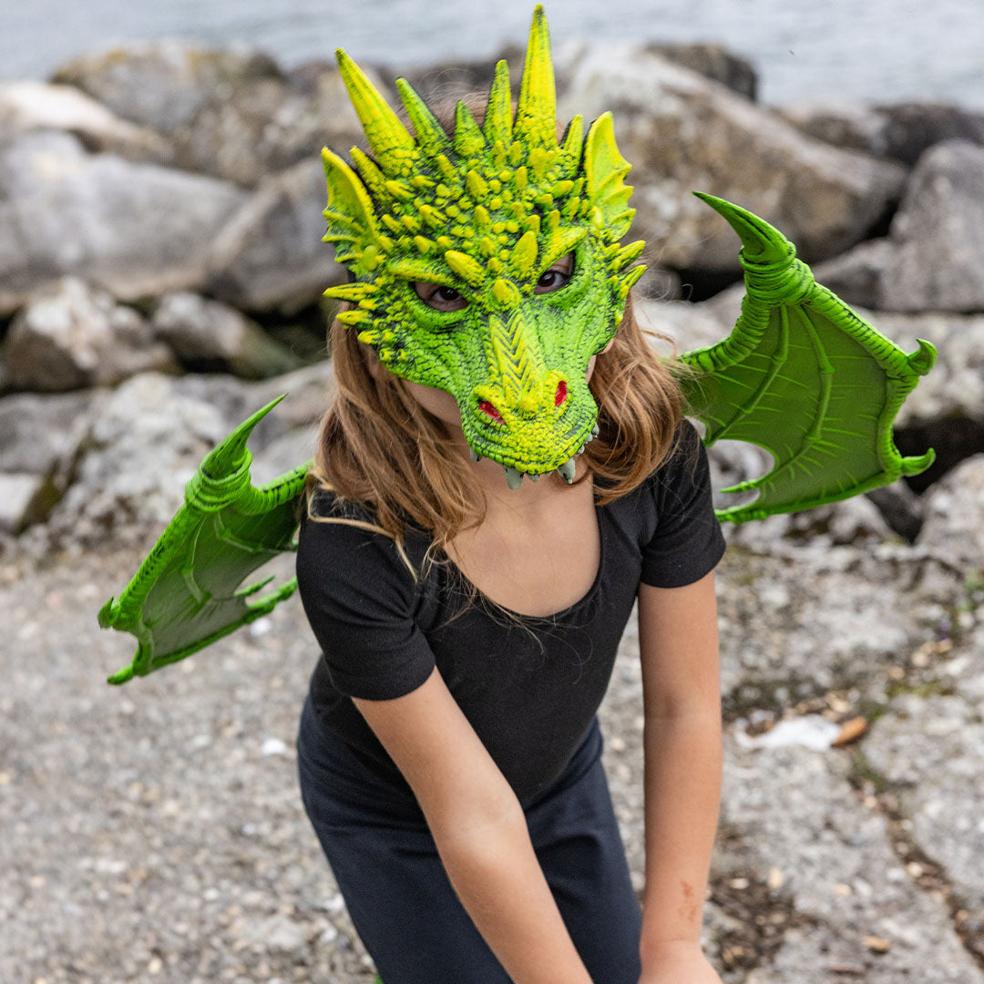 Kind draagt een groen drakenmasker en vleugels, zittend op de grond bij rotsen.