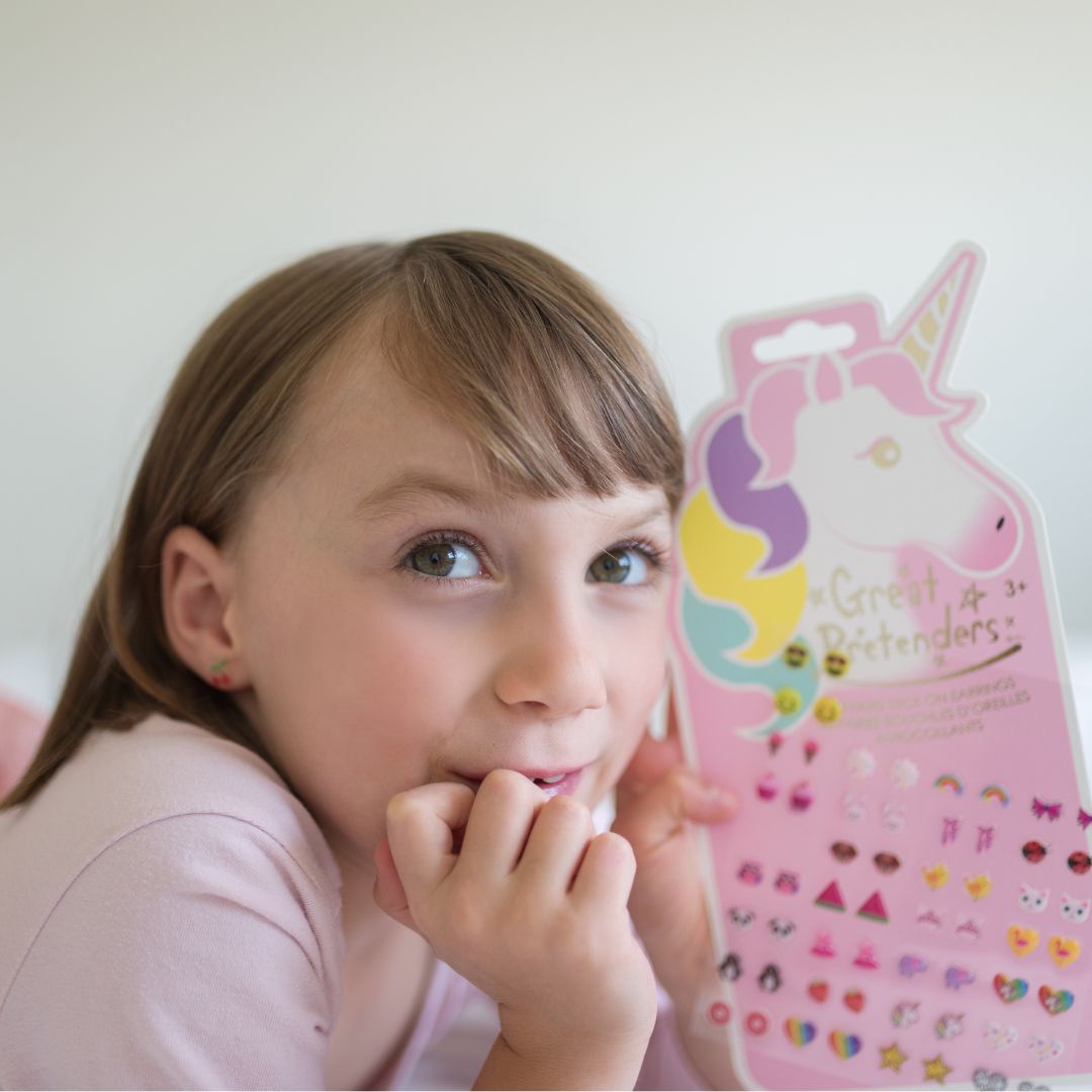 Een meisje met een kleine oorbel houdt de verpakking van Great Pretenders plakoorbellen met een eenhoorn vast.