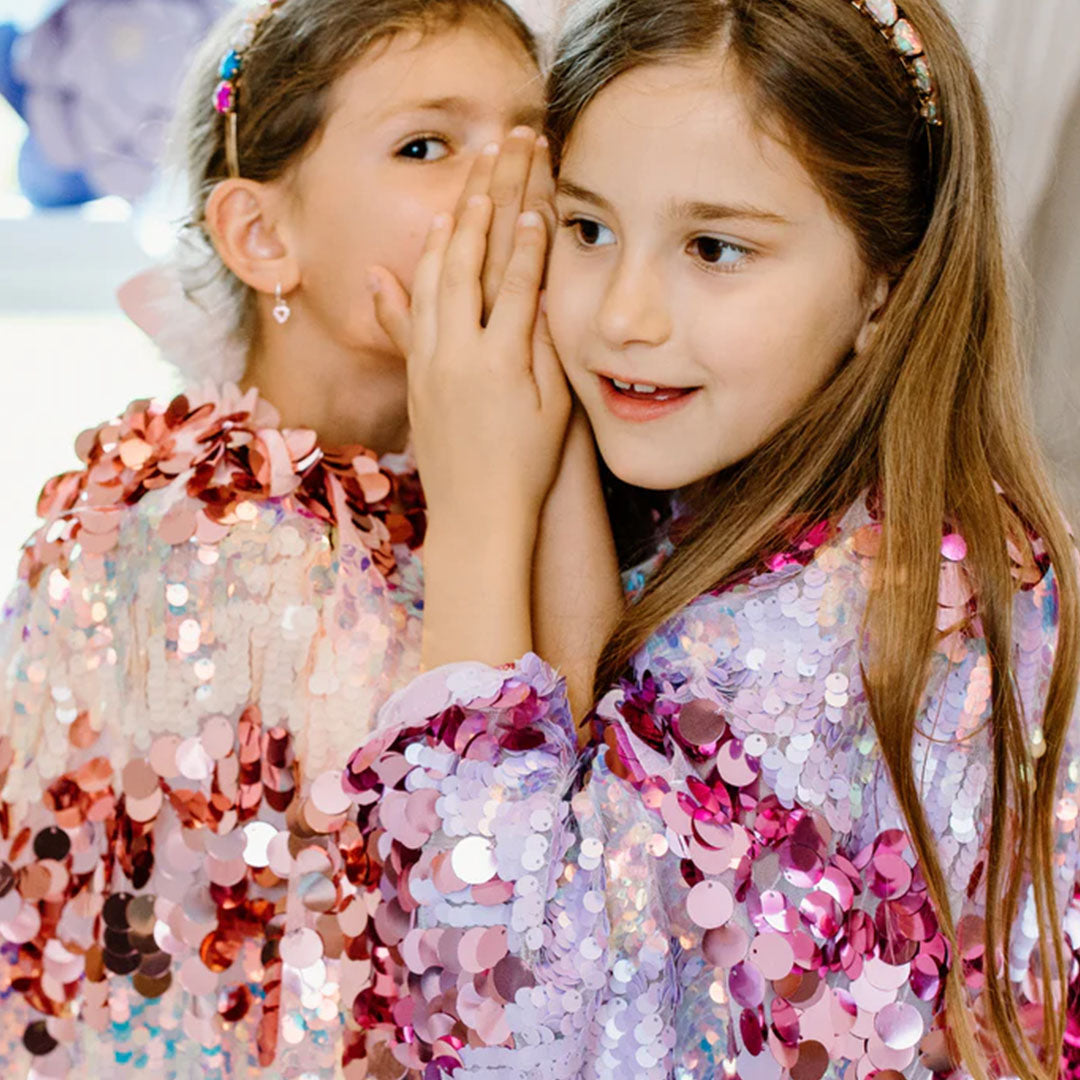 Twee meisjes, waarvan één de Great Pretenders verkleedcape voor kinderen met paarse pailletten draagt, fluisteren naar elkaar.