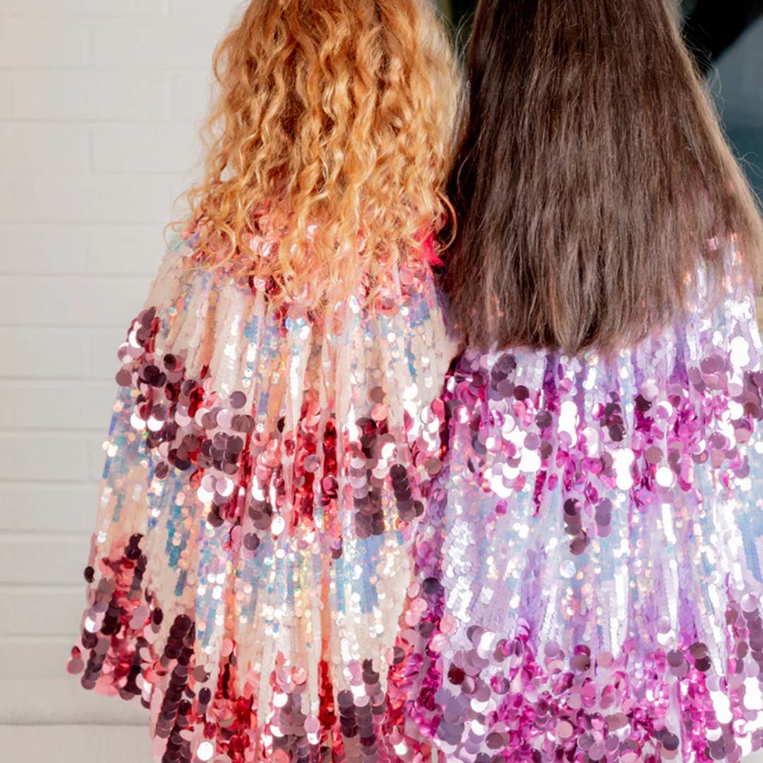 Achteraanzicht van twee meisjes die de Great Pretenders verkleedcape voor kinderen met paarse pailletten en een roze variant dragen.
