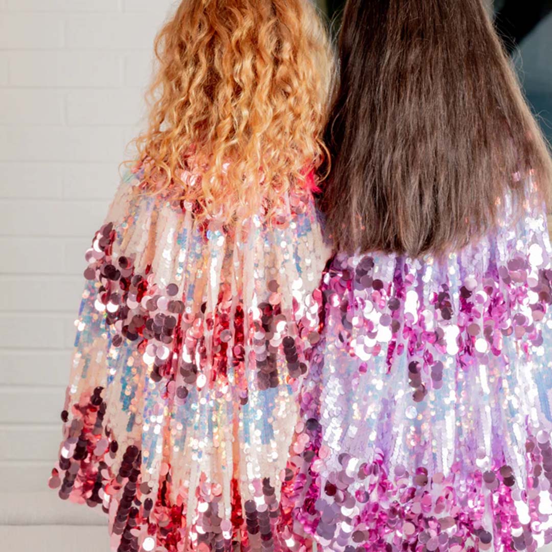 Twee meisjes dragen van achteren gezien een roze en een paarse verkleedcape met pailletten.