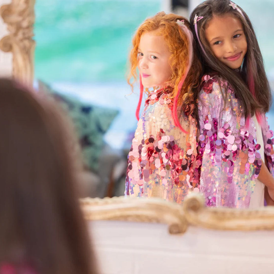 Twee meisjes met een roze en een paarse verkleedcape met pailletten kijken in een spiegel.