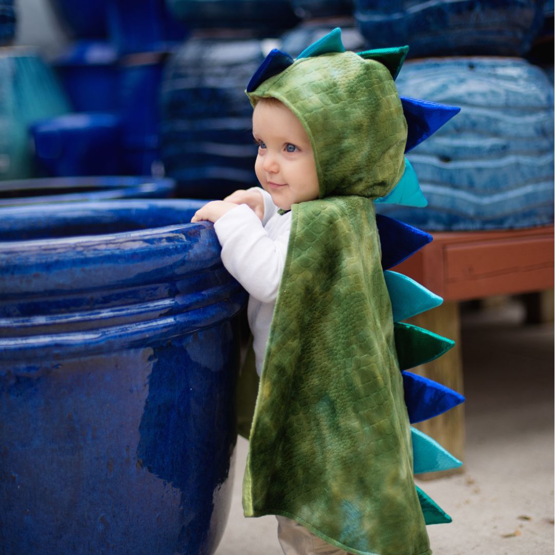 Een baby draagt de Great Pretenders groene draken verkleedcape en staat naast een grote blauwe pot.