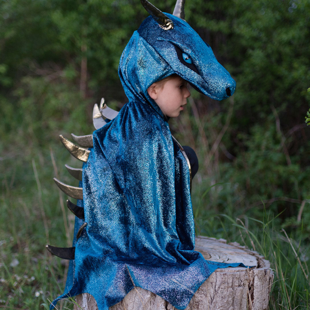 Kind draagt de blauwe Great Pretenders draken verkleedcape en zit op een boomstronk in het bos.